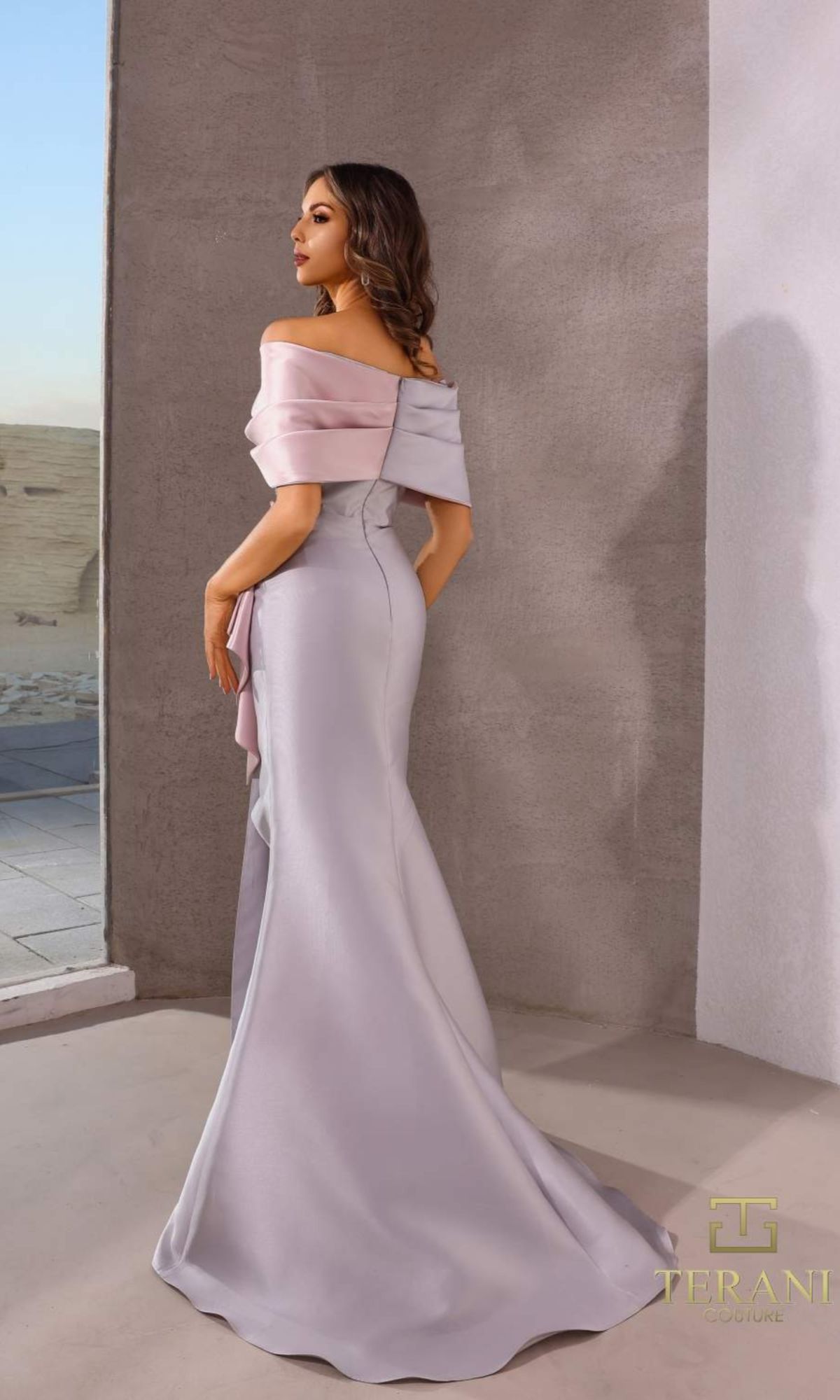 A woman stands indoors in the Terani 252M5324 long formal dress, a pale pink and lavender off-the-shoulder mermaid gown with a fitted silhouette and elegant draped detailing.